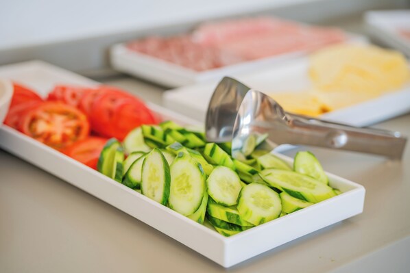 A white rectangular tray with sliced cucumbers and tomatoes, a metal spoon, and a blurred background with other food items. - Ramnus Karpathos Suites (Karpathos)