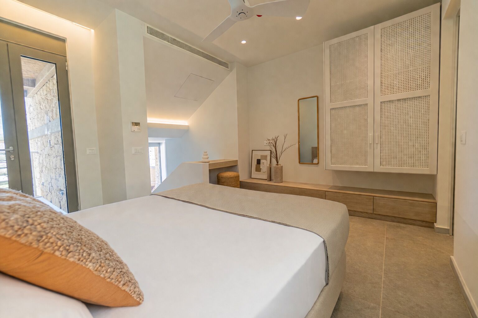A modern bedroom with a large bed, a ceiling fan, a built-in wardrobe, and a view of the outside.