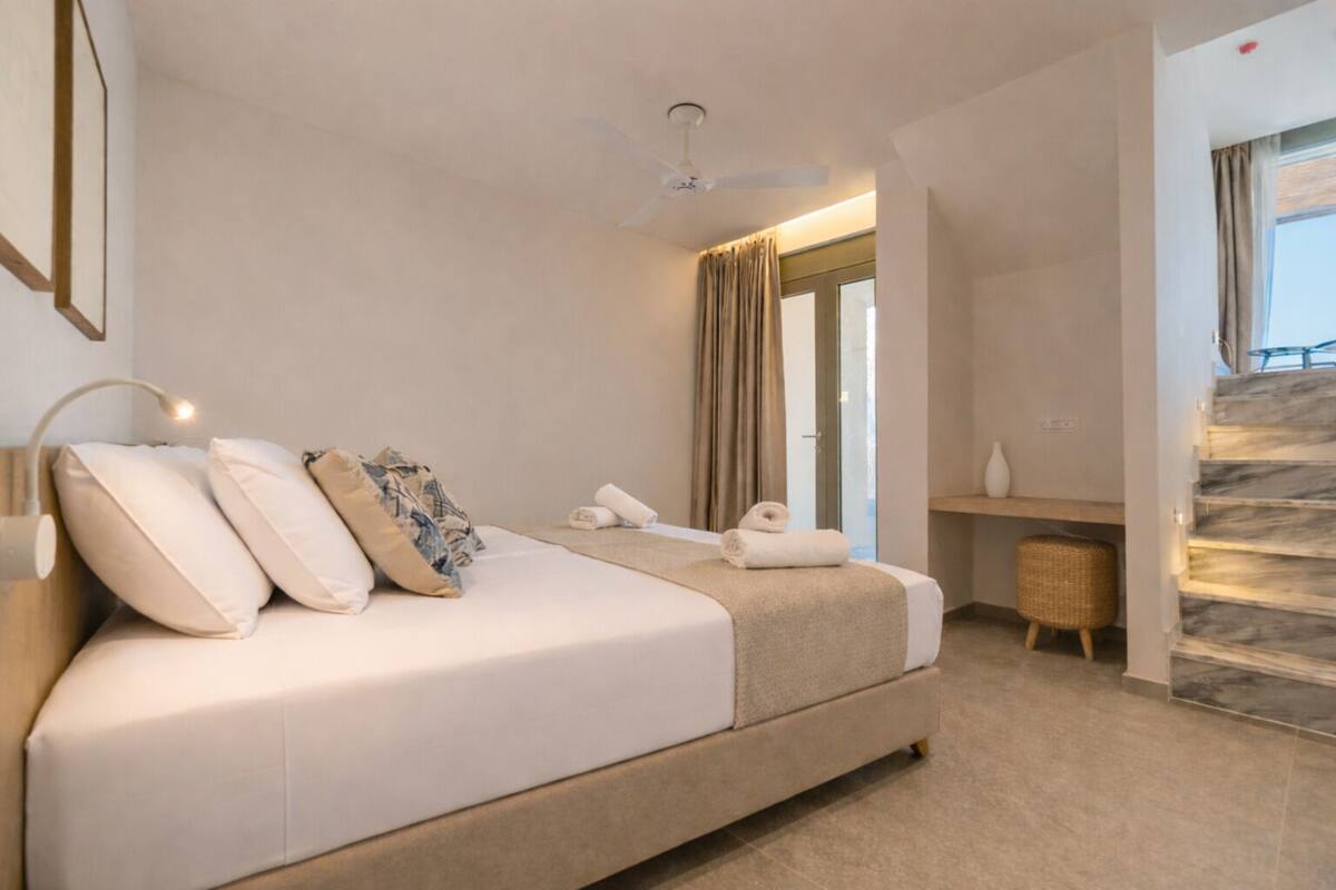 A hotel room with a bed, a ceiling fan, a window with curtains, a staircase, and a small table with a vase.