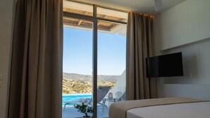 A bedroom with a bed, a TV, a potted plant, and a view of a pool and landscape. - Ramnus Karpathos Suites (Karpathos)