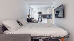 Una habitación de hotel pequeña con cama, escritorio, silla, kitchenette y televisor.