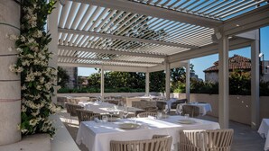 Area pranzo all'aperto con tavoli per due, coperta da un pergolato, circondata da sedie e un'elegante composizione floreale.