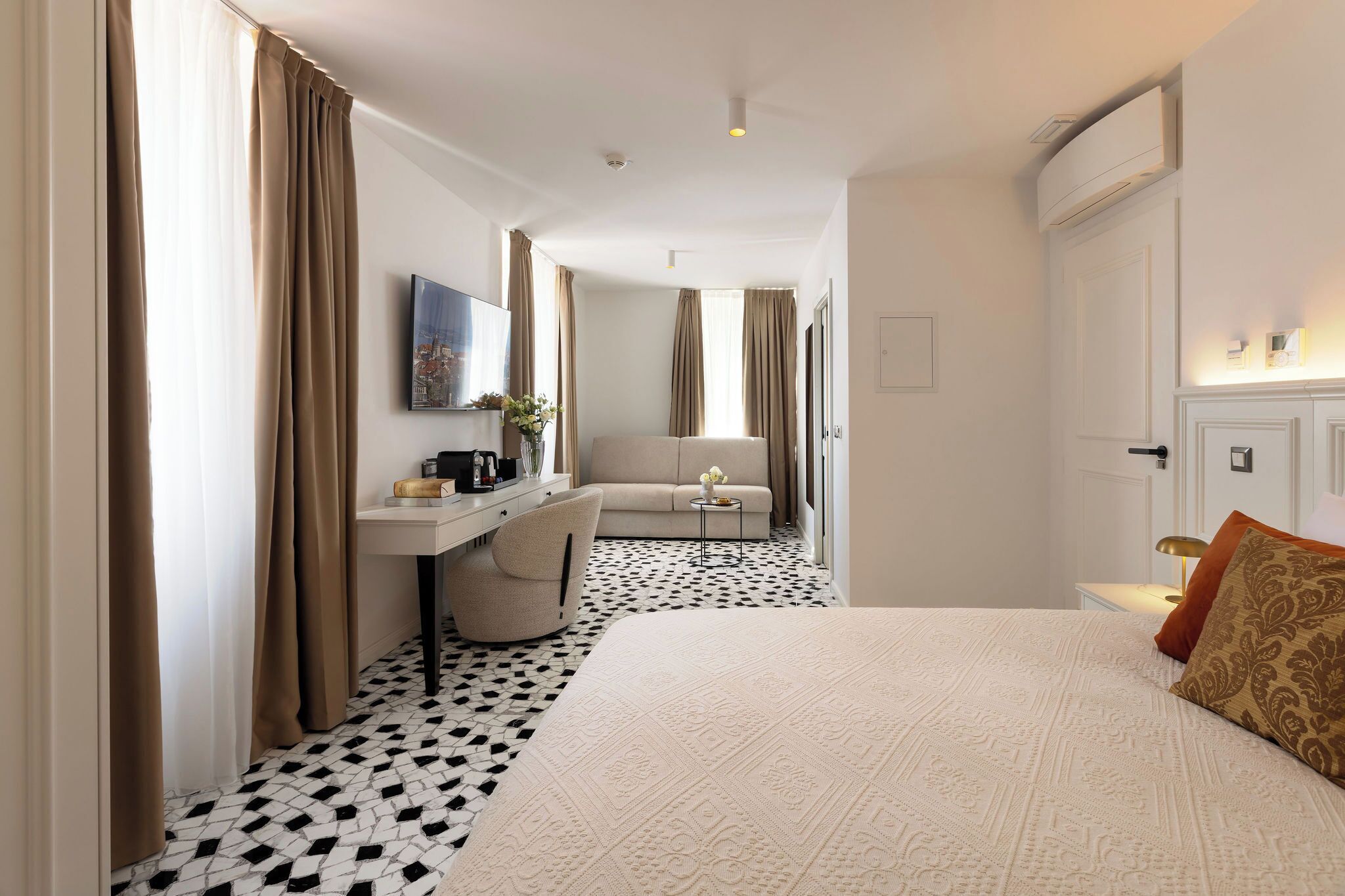 A hotel room with a bed, a desk, a chair, and a small table with a lamp.