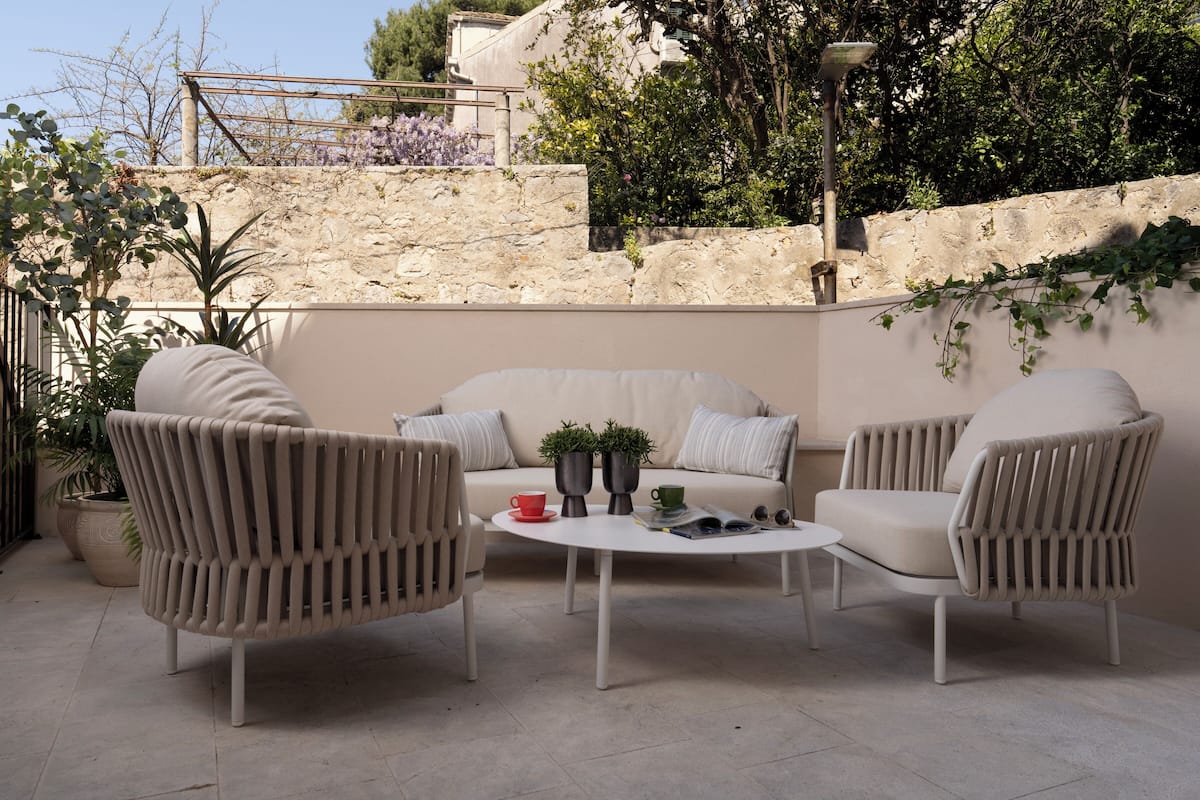 Een terras met een bank en twee stoelen, een ronde tafel, potplanten en een stenen muur op de achtergrond.