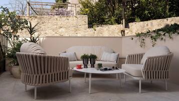 An outdoor seating area with a sofa and two chairs, a round table, potted plants, and a stone wall in the background.