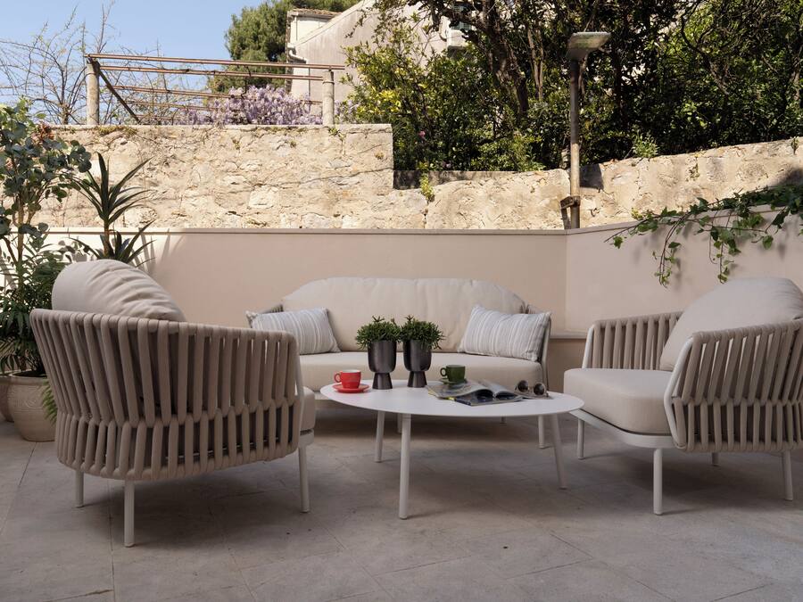 Un área de estar al aire libre con un sofá y dos sillones, una mesa redonda, plantas en macetas y un muro de piedra al fondo.