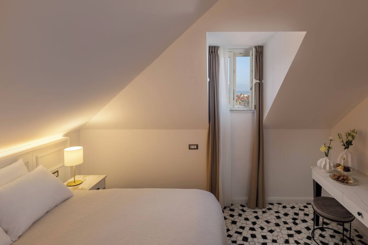Een hotelkamer met een bed, een bedlampje, een klein tafeltje met een vaas, een stoel en een raam met gordijnen.