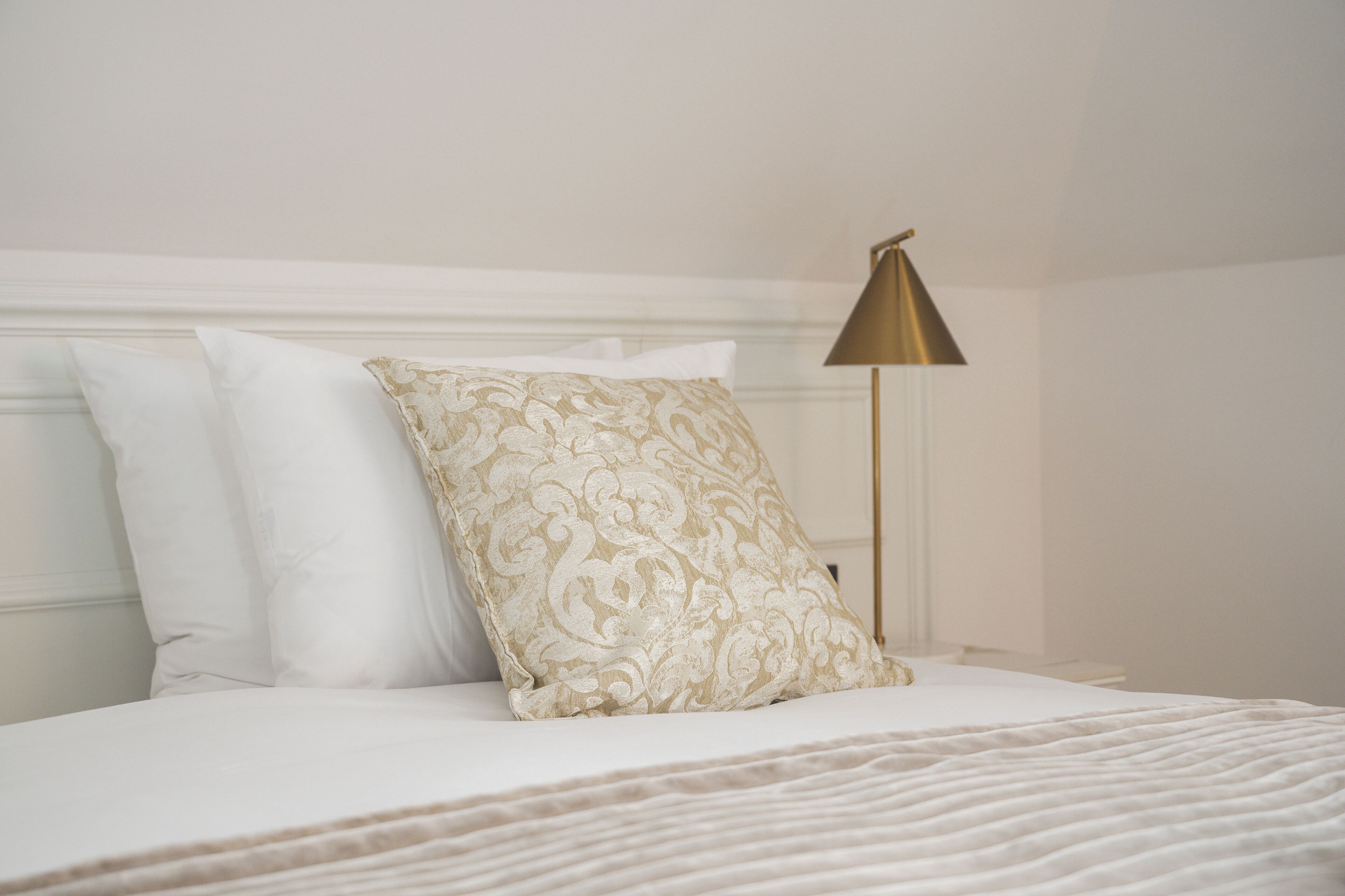 A neatly made bed with white and patterned pillows, a gold-colored lamp, and a white headboard.