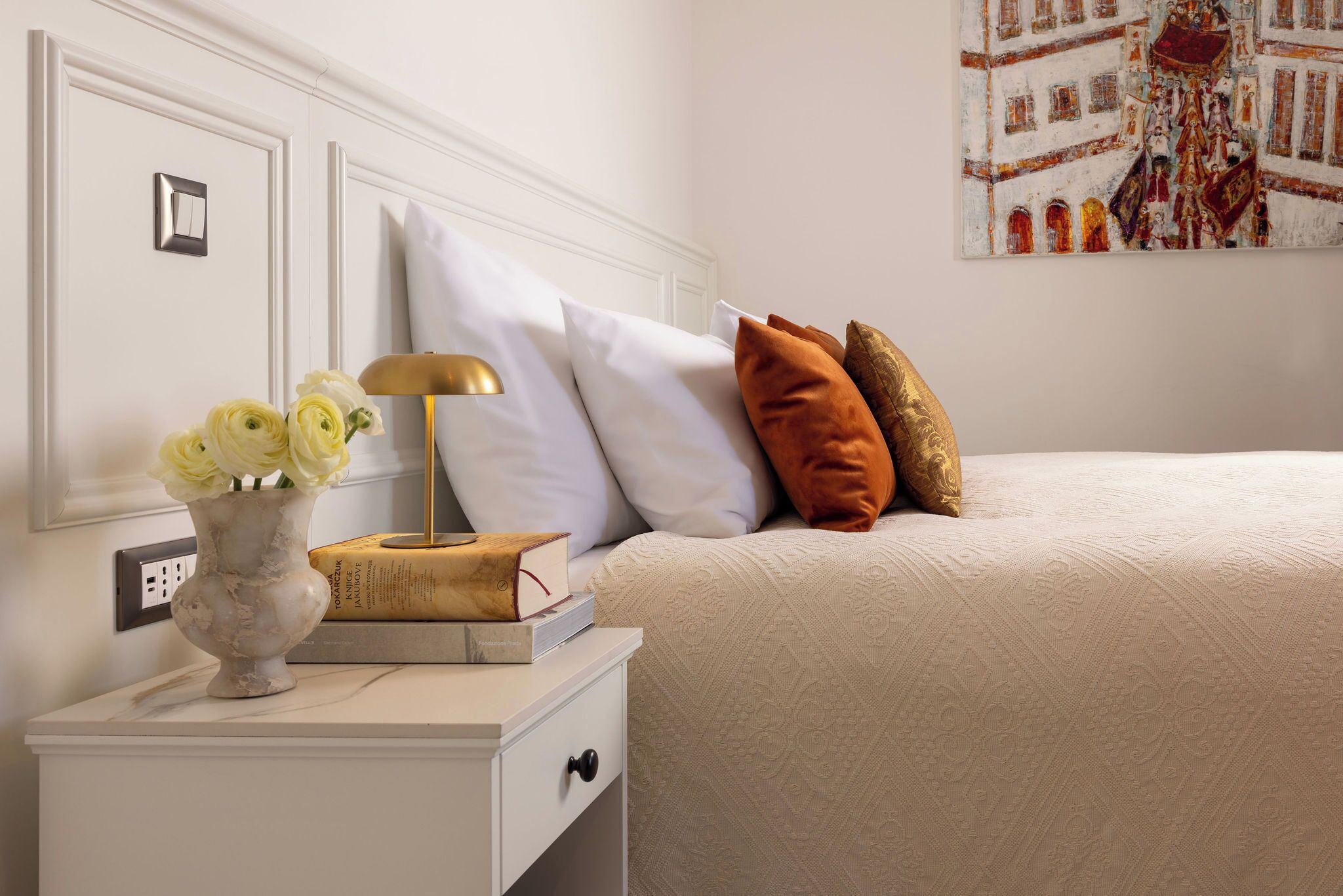 Um quarto bem organizado com uma cama, almofadas, uma mesa de cabeceira com um vaso de flores, um abajur e um livro.