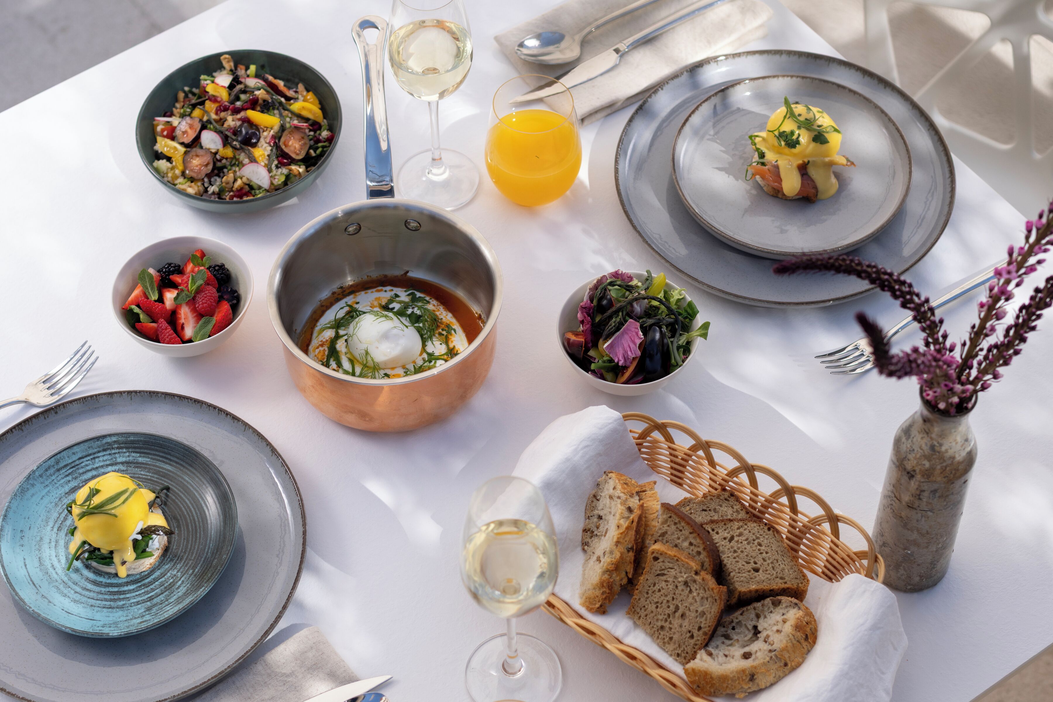 A table set with various dishes, a glass of white wine, a glass of orange juice, and a basket of bread.