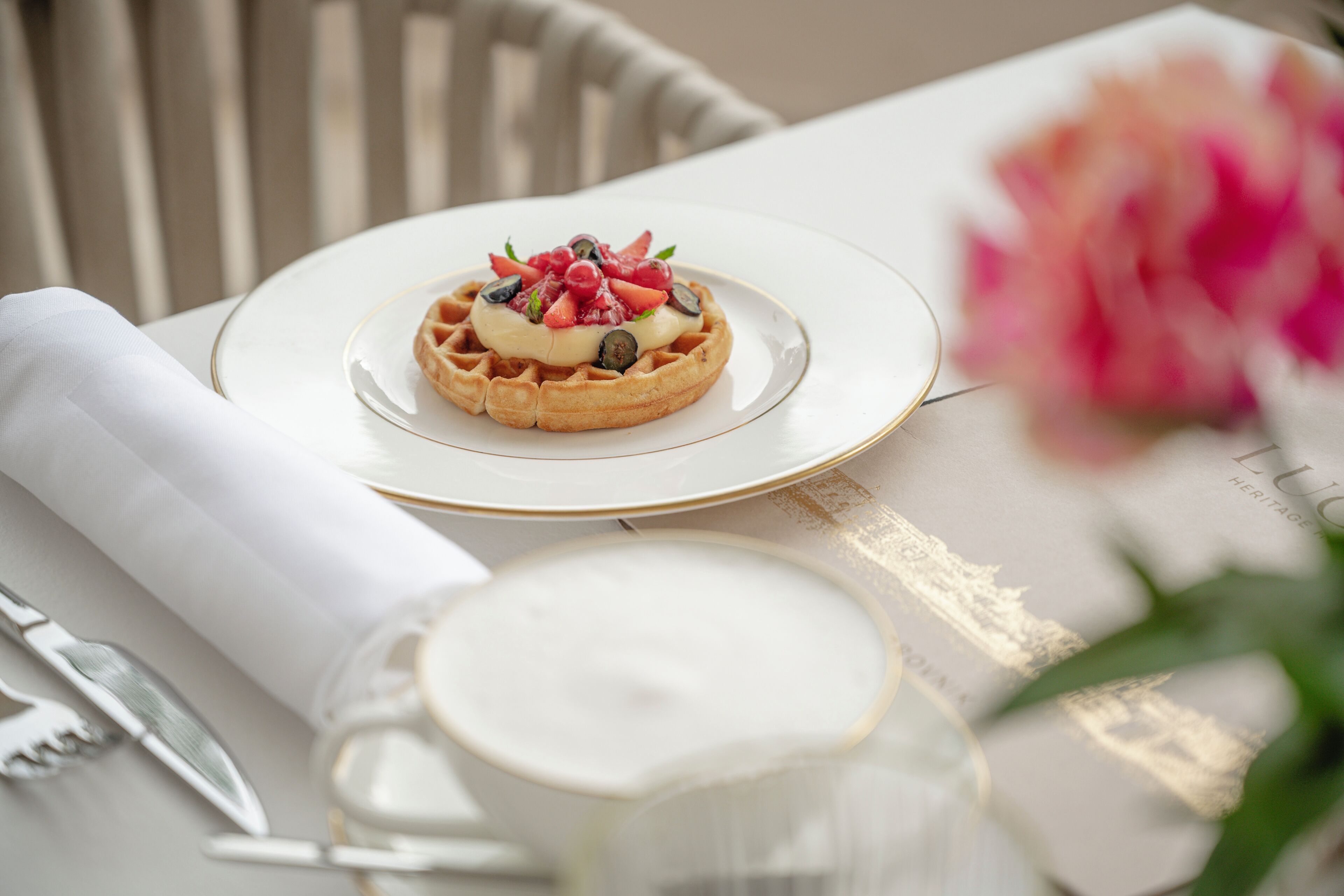 Ein Waffel mit Fruchtbeilagen auf einem Teller, ein gefaltetes Servietttuch, ein Glas Milch und ein unscharfer Blumenhintergrund.