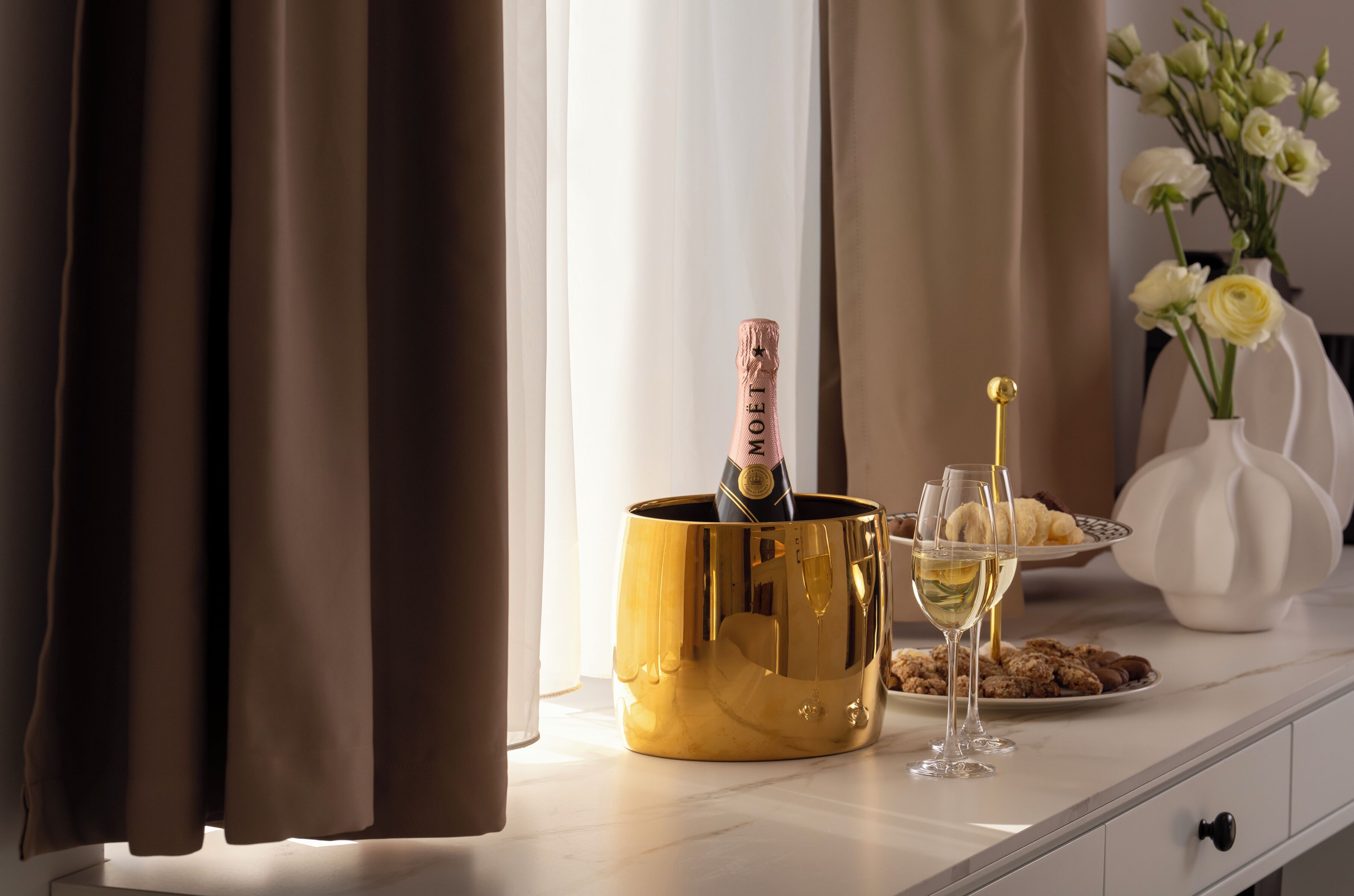 Een champagnefles in een gouden emmer, een champagneglas en een bord met koekjes op een witte tafel.