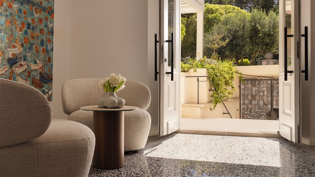 Una stanza con un divano, un tavolino con un vaso di fiori, un quadro e vista su un giardino attraverso una porta aperta.