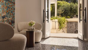 Une pièce avec un canapé, une petite table sur laquelle est posé un vase de fleurs, un tableau, et une vue sur un jardin, visible à travers une porte ouverte.