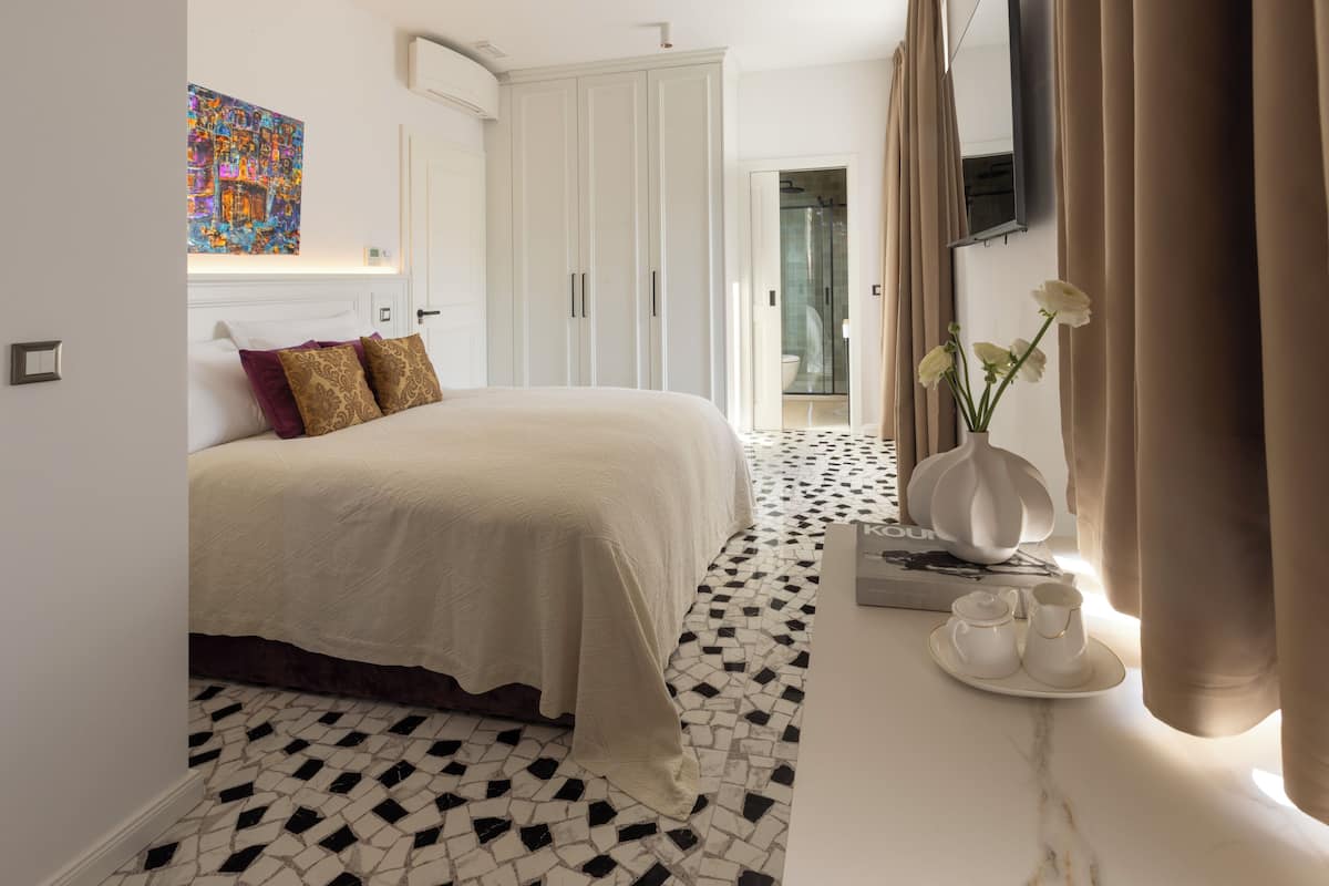 Een hotelkamer met een bed, een witte inbouwkast, een badkamer met een glazen deur, een bijzettafeltje met een vaas en kopjes, en een betegelde vloer.