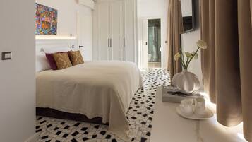 A hotel room with a bed, a white wardrobe, a bathroom with a glass door, a coffee table with a vase and cups, and a tiled floor.