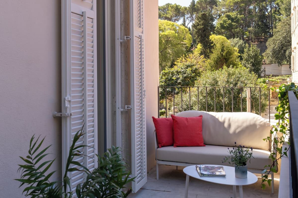 Een balkon met een beige bank, rode kussens, een klein tafeltje met een boek en een bloempot.