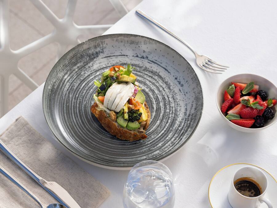 Una mesa con un plato de comida, un cuenco de fruta, una taza de café y una cesta de pan.