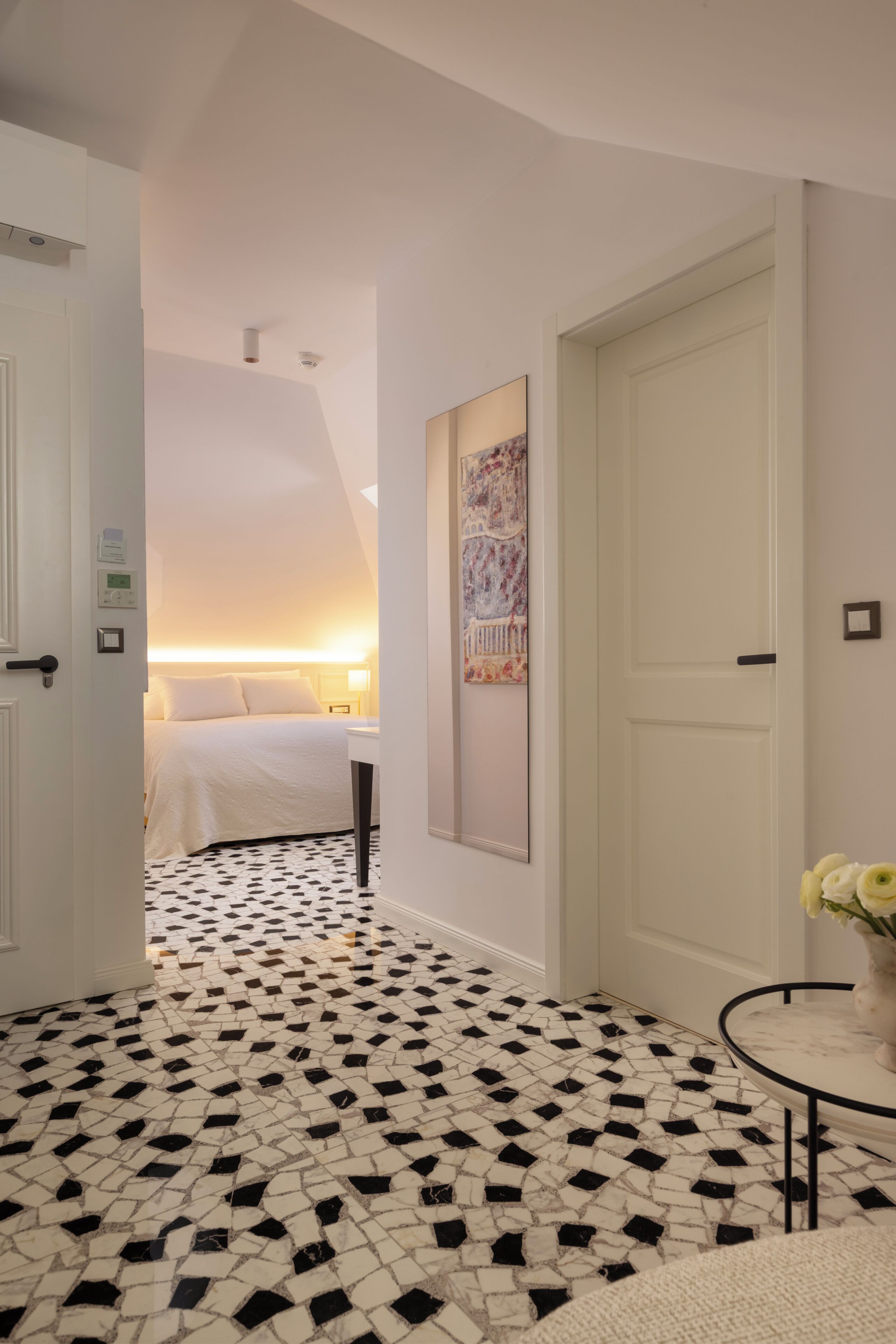 Une chambre d’hôtel avec un sol carrelé, un lit, une table de chevet, une commode, une œuvre d’art accrochée au mur et une porte.