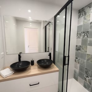 Un baño con dos lavamanos negros tipo lavabo, un espejo grande, una puerta blanca y una ducha con puertas de cristal.