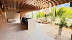 A modern hotel lobby with a reception desk, a sofa, and a large window with a view of greenery. - LM HYDE PARK (João Pessoa)