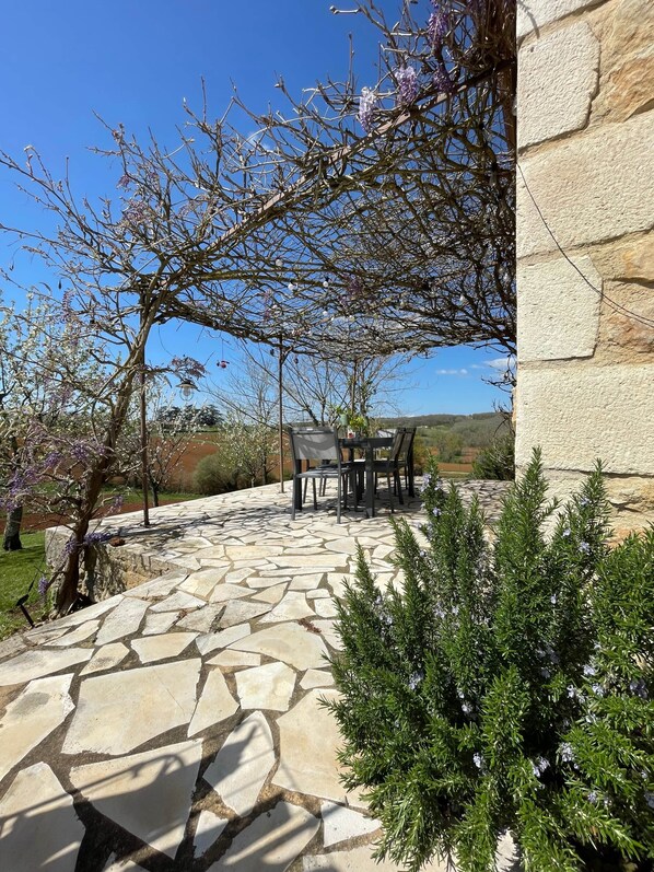 Une terrasse en pierre avec une table et des chaises, entourée de verdure et d’un mur de pierre.