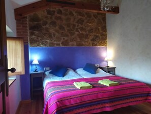 Ein Schlafzimmer mit einer Steinswand, einem Bett mit blauen Kissen, zwei Nachttischlampen und einer farbenfrohen, gestreiften Bettdecke.