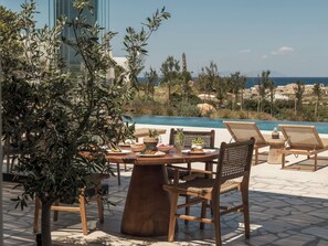 Zona junto a la piscina con una mesa redonda de madera, sillas de mimbre y vistas al mar y edificios lejanos.