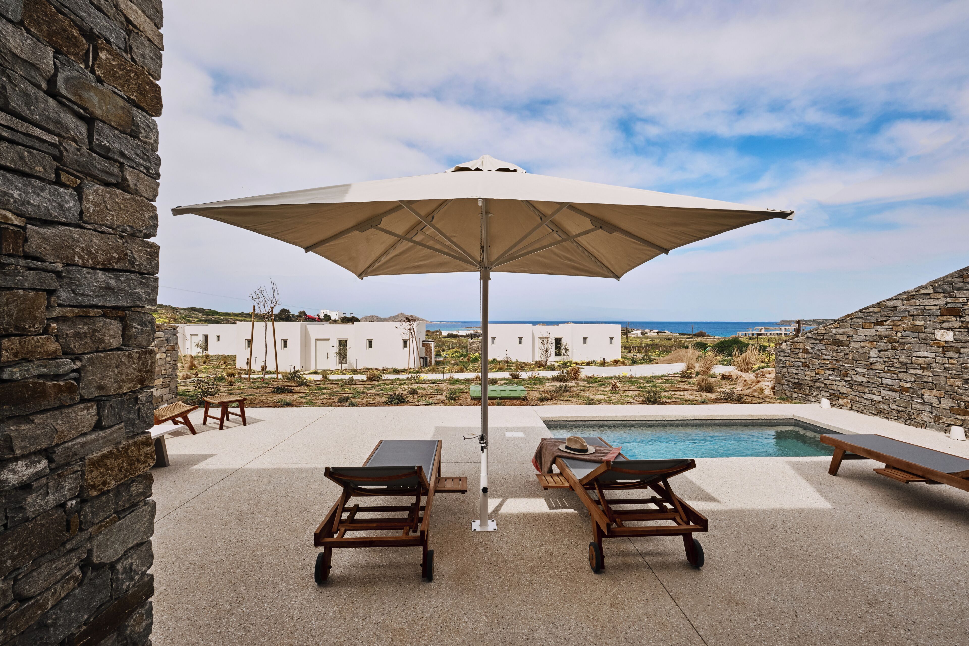 A poolside area with lounge chairs and a large umbrella, overlooking a white building and the sea.