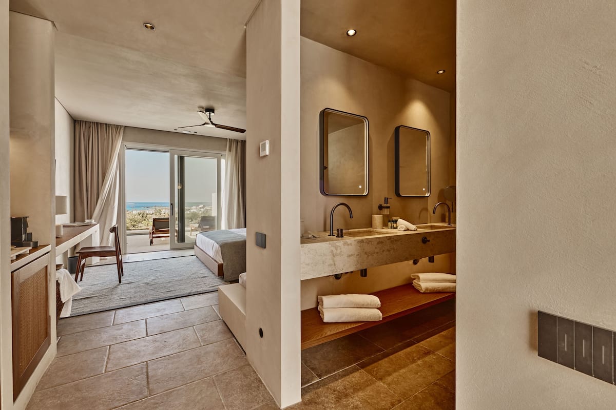 A modern hotel bathroom with a large sink, two mirrors, and a view of the sea through the glass door.