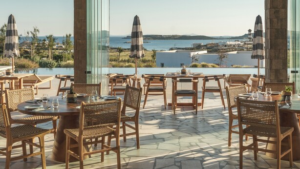 Un espace de restauration en plein air, avec des chaises et des tables en osier, offrant une vue sur un paysage côtier, la mer et des collines lointaines.