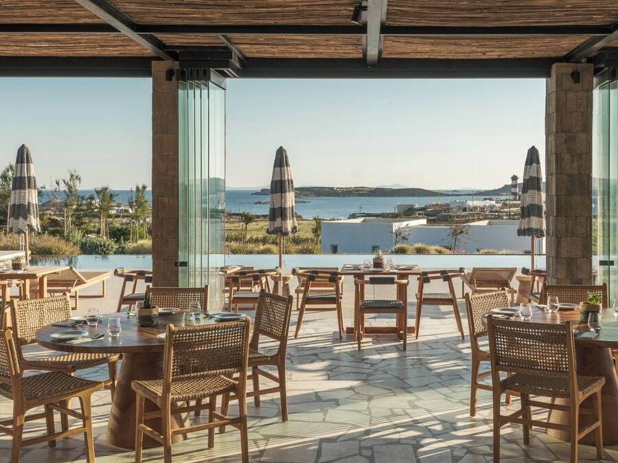 Uma área de refeições ao ar livre com cadeiras e mesas de vime, com vista para uma paisagem costeira que inclui o mar e colinas ao longe.