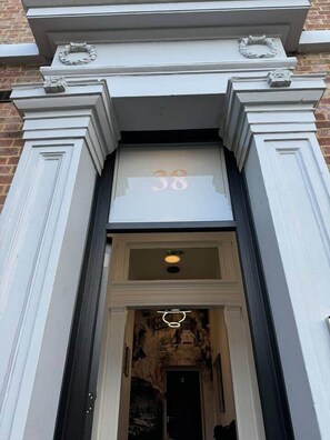 Ein Gebäudeeingang mit der Hausnummer 38 über der Tür, eine schwarze Tür und ein kleines Fenster darüber.
