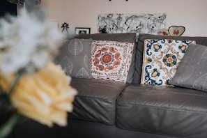 Un sofá de cuero marrón con cojines estampados, una pared con obras de arte enmarcadas, y un jarrón con flores en primer plano.