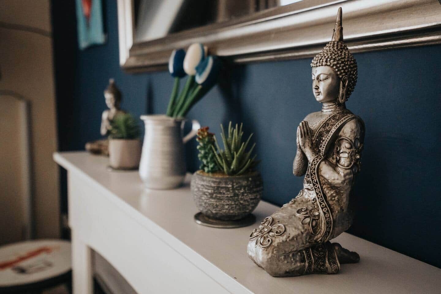 En Buddha-figur på en hvid hylde med krukker med planter og en lille skulptur.