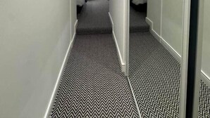 A narrow hallway with a black and white chevron-patterned floor leading to a room with a bed, a small table, and a wall-mounted light fixture.