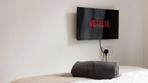 Ein Bett mit weißer Decke und einem dunkelgrauen Kopfkissen, ein schwarzer, an der Wand montierter Fernseher mit dem Netflix-Logo und eine Steckdose an der Wand.
