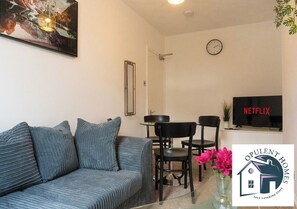 A living room with a sofa, a dining table with chairs, a TV, and a clock on the wall. - Delightful 5-bedroom house in charming Reading perfect for families (Reading)
