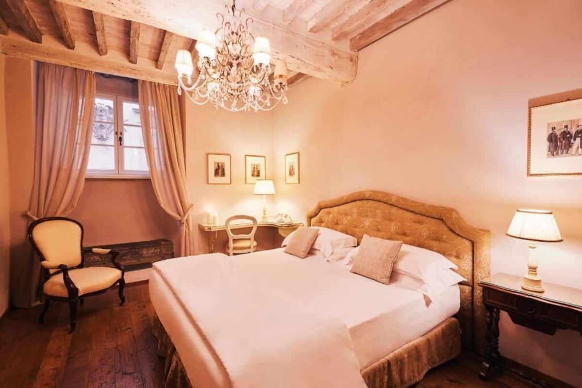 A bedroom with a large bed, a chandelier, a desk with a lamp, and a chair.