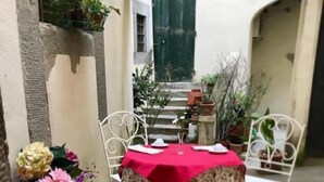 Ein schmales Innenhof mit einem für zwei Personen gedecktem Tisch, umgeben von Topfpflanzen und einer roten Tischdecke.