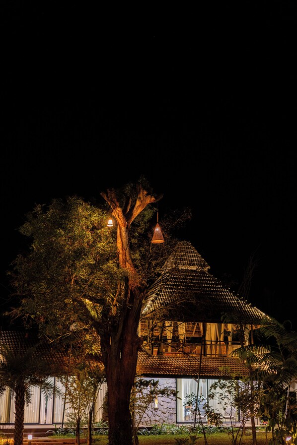 Un bâtiment traditionnel avec un toit en tuiles, entouré d’une végétation luxuriante et d’un grand arbre.