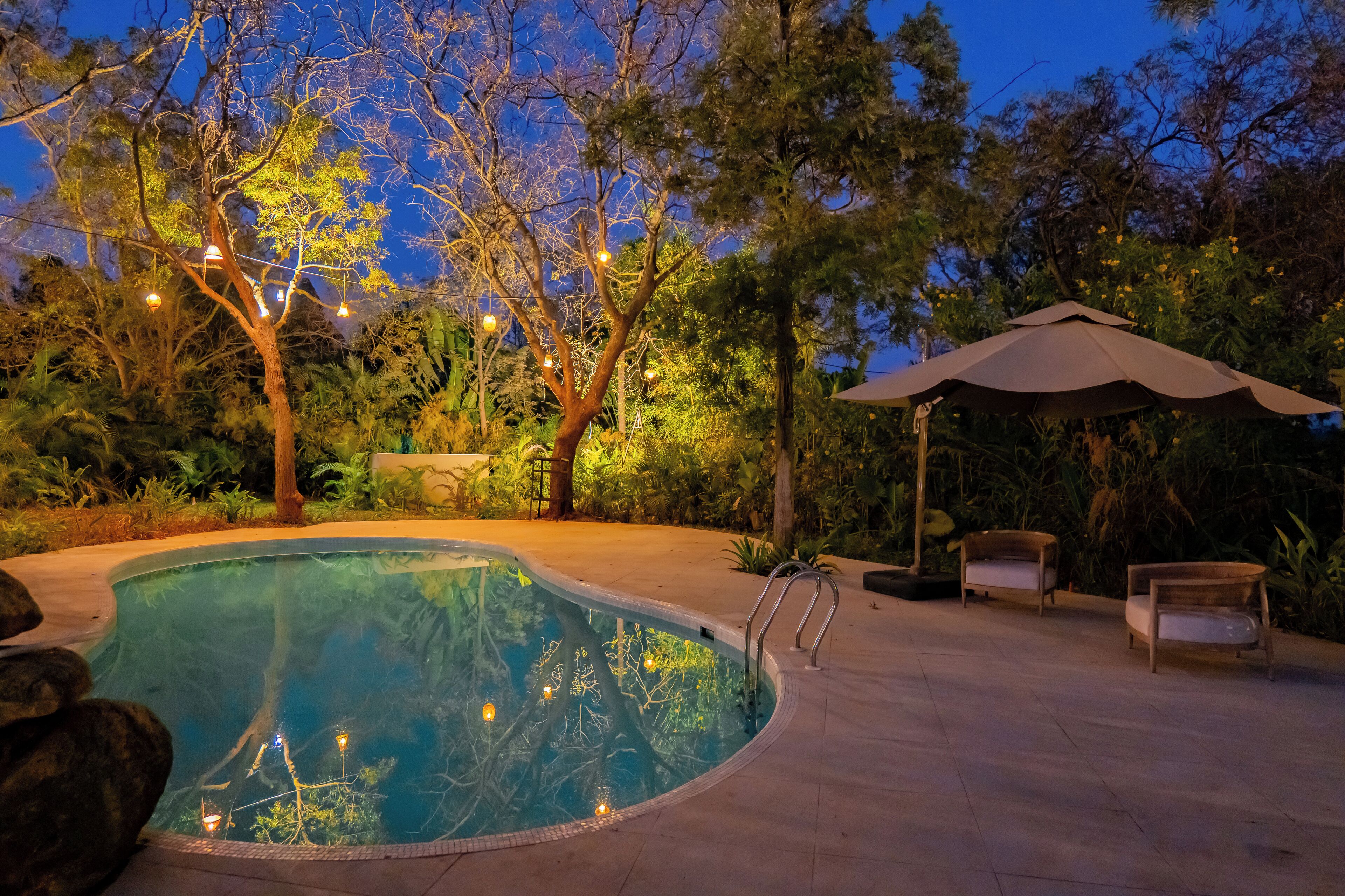 A swimming pool with a tree in the center, surrounded by patio furniture and an umbrella.