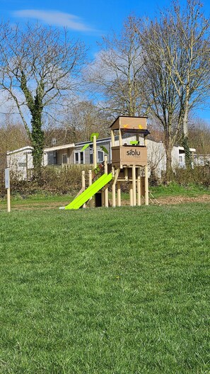 Un parque infantil con tobogán y estructuras para escalar, rodeado de árboles y un campo cubierto de césped.