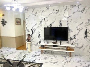A living room with a marble-patterned wall, a flat-screen TV, a white entertainment unit, and a decorative bust. - PLOT 1792 LUXURY SUITES (Abuja)