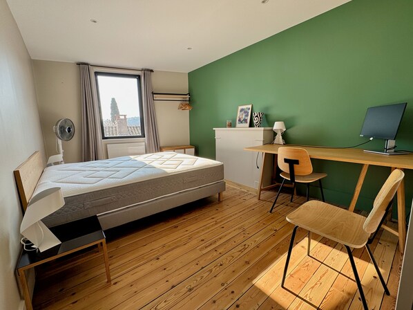 A bedroom with a bed, a desk, a chair, a window, and a view of a building.