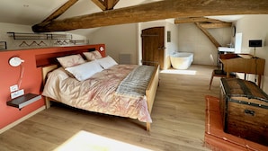 Camera da letto con soffitto in legno, un letto con biancheria a motivi, un baule in legno, una vasca da bagno e una piccola scrivania con una lampada.