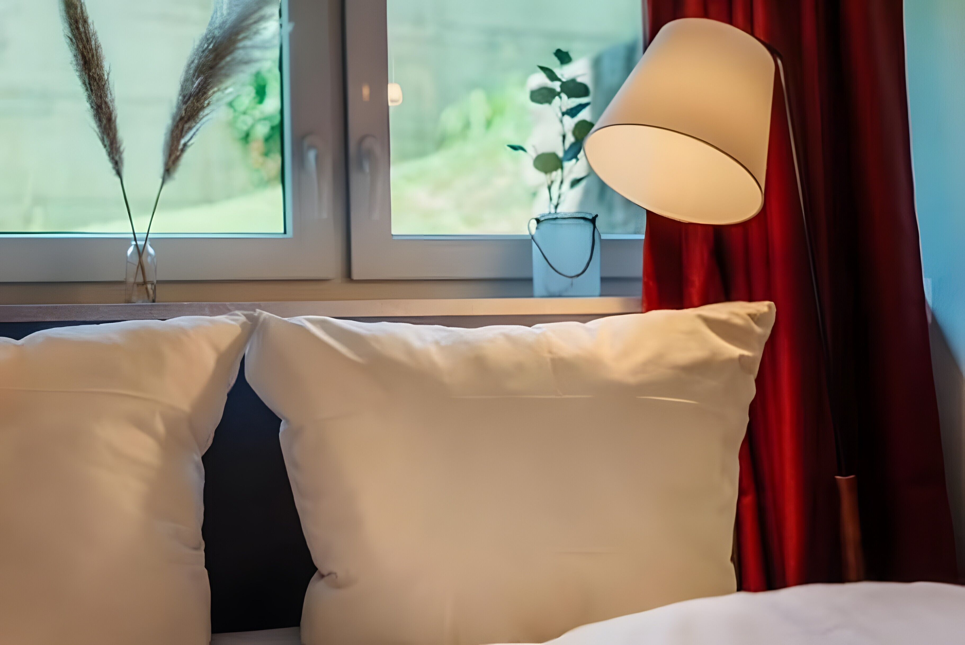 A bed with white pillows, a bedside lamp, a window with a view of greenery, and a vase with pampas grass.
