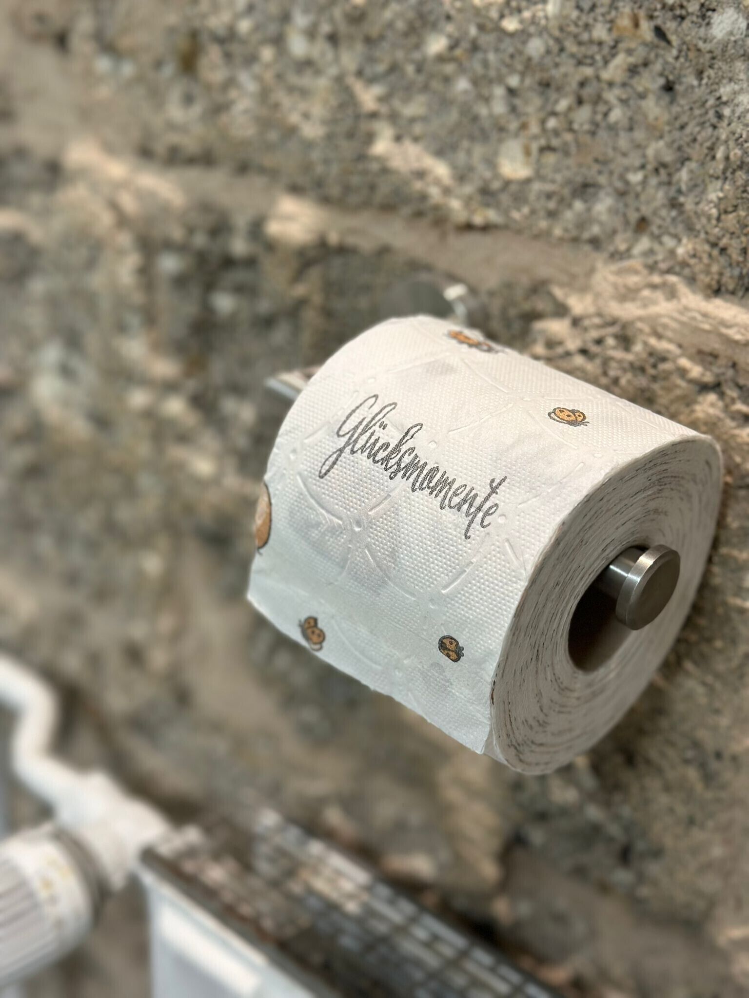 Un rouleau de papier toilette portant l’inscription « Glücksmanufaktur », fixé sur un mur à texture.