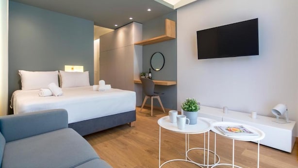 A modern hotel room with a bed, a desk, a chair, a small table with two cups, and a TV mounted on the wall. - Athena Urban Retreat Apartment (Athens)