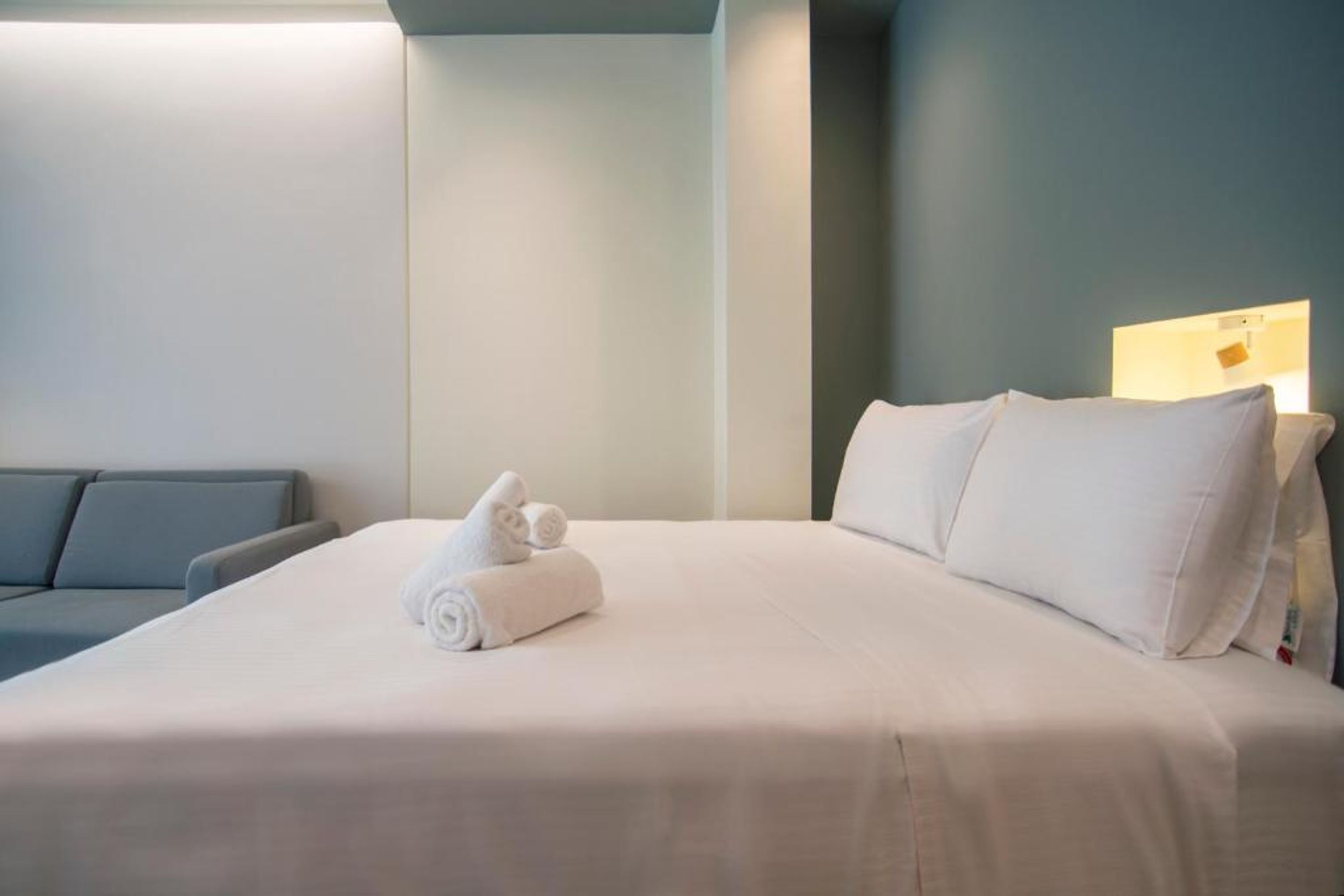 Een net opgemaakt bed met witte lakens en een opgerolde handdoek, een grijze bank en een wandlamp.